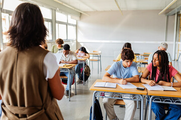 High school students writing during exam with teacher supervising. Focused group of young learners...