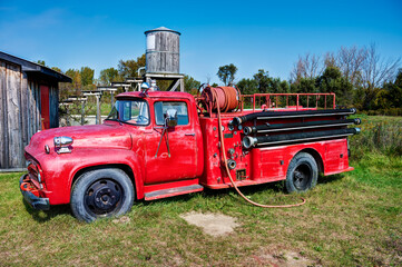 Close-up of a vintage red fire truck parked on grass beside a rustic wooden building, with firefighting hoses and equipment under a bright blue sky in a rural countryside setting.