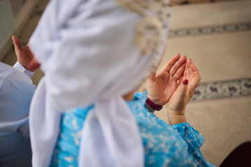 Obraz premium Closeup hands raised in prayer young girl wearing blue hijab kneeling on patterned carpet inside mosque delicate bracelet warm light serene focus spiritual devotion