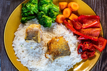A perfectly roasted fish fillet served on a plate with steamed rice, broccoli florets, and sliced carrots. The golden-brown fish contrasts beautifully with the vibrant green broccoli and bright orange
