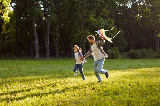 Happy father and child holding hands and running together on sunny field while flying colorful kite. Joyful family enjoying outdoor leisure and playful activity in green park surrounded by trees.