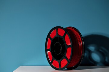 Red filament spool for 3D printing sits on a white surface against a blue background, showcasing vibrant color and design for creative projects and innovation