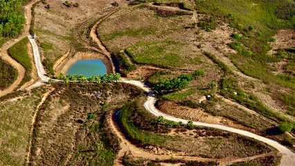 Aerial view of a dry hilly landscape with winding dirt roads, a small turquoise pond, and patches...