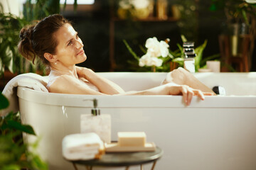 A woman enjoys serene relaxation in a white freestanding bubble bath, a peaceful smile. Lush green plants, orchids, & warm lighting create a luxurious home spa environment for self-care.