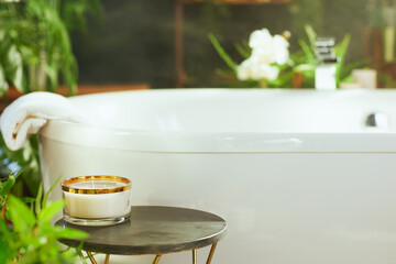Tranquil bathroom with white bathtub & rolled towel. Lit candle on table with lush green plants...