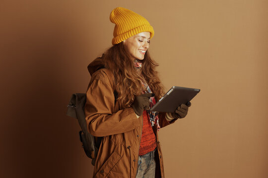 A smiling woman with a yellow beanie and brown jacket points at a tablet screen, likely planning or sharing information for an outdoor adventure or travel.