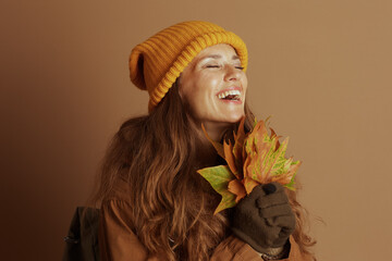 A woman, wearing a yellow beanie and brown jacket, laughs with her eyes closed while holding a fan...