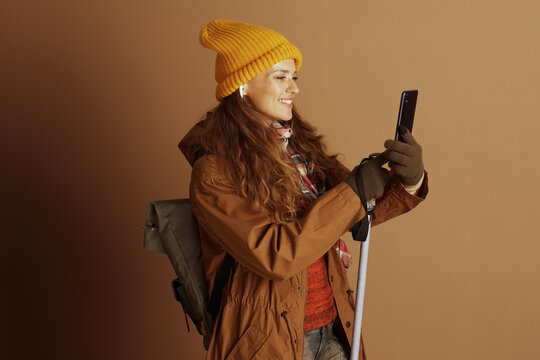 A cheerful woman in autumn hiking gear, including a yellow beanie and green gloves, smiles while checking her smartphone. She holds a trekking pole and wears a backpack, ready for outdoor adventure.
