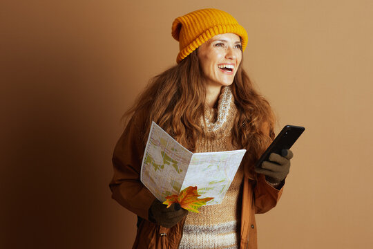 With a joyful smile, a woman in a yellow beanie and brown jacket holds a map and smartphone, looking right, embodying the excitement of an upcoming autumn hike or travel adventure.