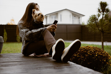 A young woman relaxing on a wooden terrace, backyard patio with her Yorkshire terrier doggy while using a smartphone. A small dog, enjoying the outdoors while looking at phone. Suburban lifestyle.