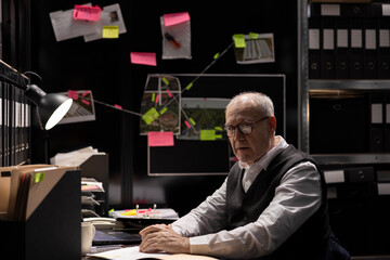Senior private detective studying photos and documents in incident room, tracing suspect activity during a clandestine operation to ensure justice and uncover organized crime. Law agency.