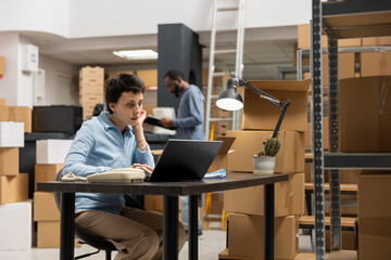 Small business employee oversees order processing at a desk in industrial depot, handling packages...