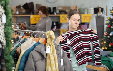 Fototapeta premium In sales area of store during Christmas sale, teen girl customer chooses coat or jacket. Creating capsule wardrobe, branded clothing at affordable prices. Modern urban fashion, casual practical