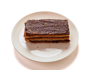 A dessert layered cake with chocolate is on a white plate on white background. The dessert is a cake with a brown crust. The plate is white and the dessert is the main focus of the image