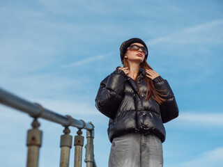 Obraz premium Young teenager girl in black jacket and grey jeans is standing on top of a stair with metal barrier, blue cloudy background. Model is wearing hat and sunglasses and enjoys warm sunny winter day.