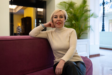 A blonde woman in a knitted sweater is sitting on a sofa in a modern interior. She is wearing a white sweater and black trousers.