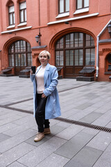 A blonde woman in a white sweater and scarf stands outside a business building. The architecture of the building is dominated by red brick.