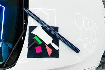 Roll of purple chameleon tint film and tools are prepared for windshield installation inside a detailing studio. Iridescent purple tint film and installation tools on car hood in detailing workshop.