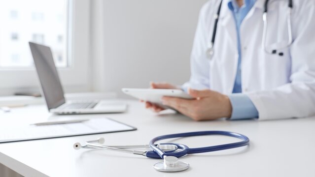 Closeup of blue stethoscope is lying on the table near female doctor reviewing prescription on digital tablet, representing digital healthcare workflow in contemporary medical setting. Medicine