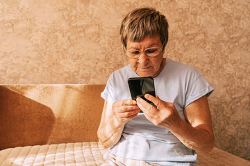 An elderly woman wearing glasses was intently immersed in her smartphone while sitting on a sofa in a sun-drenched. Digital Phone Addiction in Older Women, Screen Time Overload, and Modern Technology