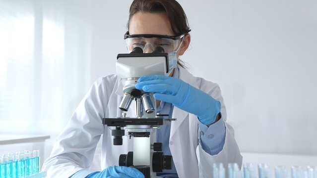Female scientist examining microscope slide, wearing protective gear in sterile laboratory, researching pharmaceutical breakthrough with blue tinted test tube specimens