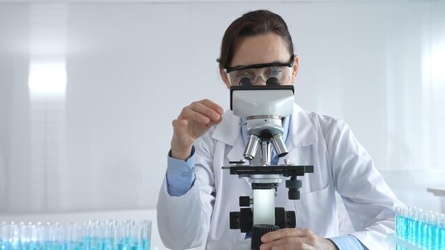 Female scientist wearing lab coat and protective glasses using microscope, analyzing samples in test tubes in modern laboratory, conducting scientific research - Powered by Adobe