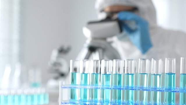 Male medical scientist in protective suit using microscope while analyzing samples, conducting research in laboratory. Close-up of test tubes with blue liquid