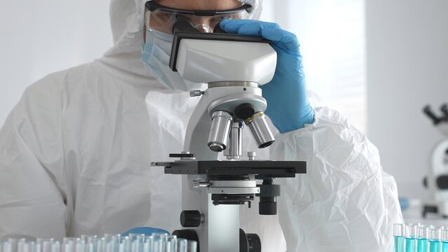 A medical virologist in protective gear and blue gloves examines viral samples under a microscope while conducting research in a sterile laboratory setting