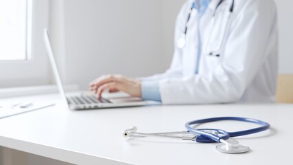 Closeup of blue stethoscope is lying on the table near female doctor working on laptop, highlighting digital tools transforming healthcare diagnostics and patient documentation. Medicine concept