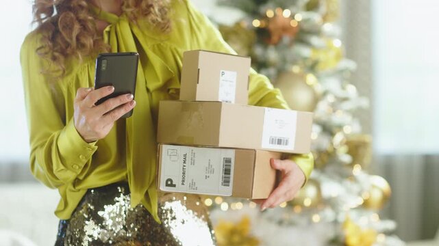 A stylish woman with curly blonde hair, in green satin blouse and sequined skirt, holds stack of shipping boxes and smartphone.