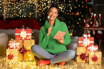 Happy thoughtful woman sitting on floor among wrapped gift boxes and garland lights, thinking and writing letter to Santa Claus