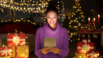 Happy lady sitting in decorated room with Xmas gifts and garlands, using digital tablet, spending...