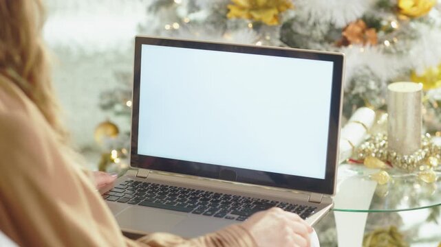 A woman with long blonde hair types on laptop with blank screen, immersed in cozy, festive Christmas setting. A beautifully decorated white Christmas tree with golden lights glows softly in background