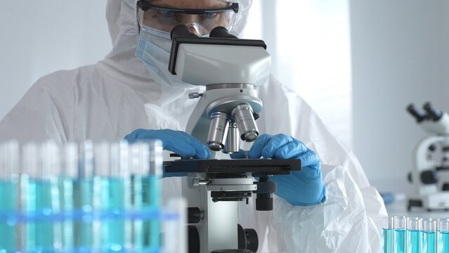 A male doctor in a protective suit and gloves uses a microscope while analyzing samples, conducting research in a laboratory. Close-up of test tubes with blue liquid - Powered by Adobe