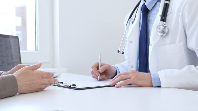 Doctor man taking notes while listening to a female patient explaining her symptoms during a medical consultation in the clinic office. Medicine and health care concept