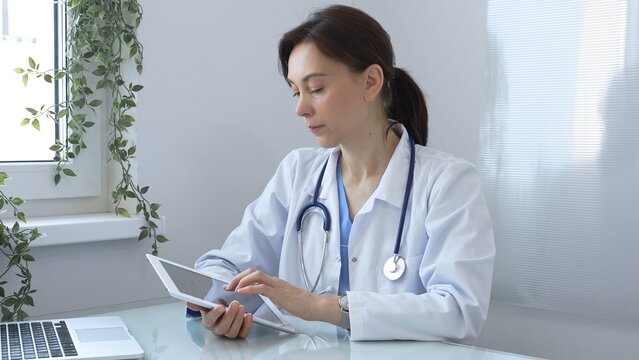 Confident female doctor wearing lab coat and stethoscope using digital tablet while sitting at desk in medical office, browsing patient information or conducting online consultation. Medicine concept