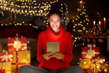 Satisfied woman using digital tablet, sitting in decorated room with Xmas gifts and garlands,...