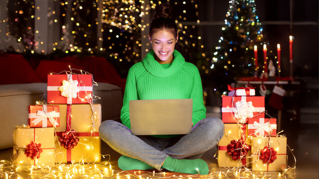 Excited woman using laptop in decorated room among gift boxes and festive lights in the evening, buying and ordering Xmas gifts