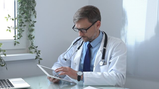 Medical doctor wearing lab coat and stethoscope using digital tablet consulting patient data in bright medical office with laptop next to him. Medicine and health care