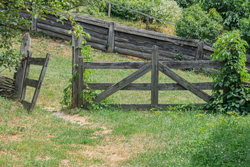 Rustic rural scene, old weathered wooden fence with ajar gate, grassy hillside, dry patchy green grass, midday lighting, no human presence, muted colors, realistic style, low angle perspective