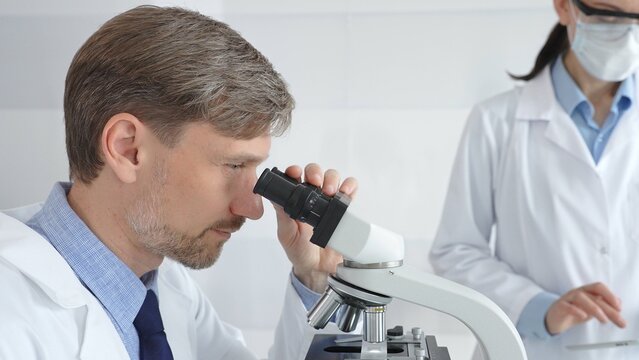 A male researcher examines a sample under a microscope, a female lab technician in a protective mask stands nearby, both in white lab coats during the study. Medicine and Science