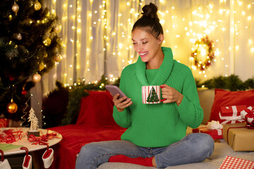 Cozy winter evening. Lady using cellphone and drinking tea or cocoa, enjoy festive holiday mood,...