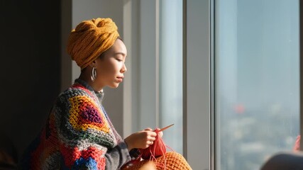 A serene young woman in a vibrant headscarf and handmade crochet shawl knits by a bright window. Natural light highlights her calm focus and artistic expression in a cozy modern space