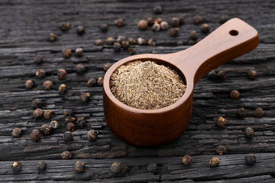 Ground black pepper in a spoon surrounded by whole peppercorns on a wood background