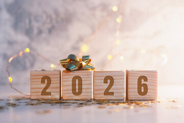 Minimal New Year 2026 composition with wooden cubes showing the year, decorated with a golden ribbon bow and warm fairy lights. Festive background for holiday greeting cards, banners and calendars.