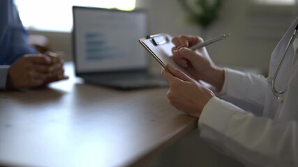 A doctor is writing on a clipboard in front of a laptop. The doctor is wearing a white coat and is talking to a patient - Powered by Adobe