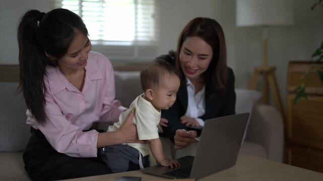 Three women are sitting on a couch with a baby in between them. One of the women is holding the baby while the other two women are looking at a laptop. Scene is warm and friendly