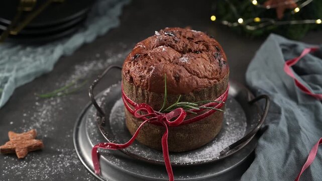 A delicious, rustic Panettone cake is wrapped in brown paper and tied with a red ribbon and green rosemary sprig, centered on stacked metal platters against a dark background. The scene is dusted with