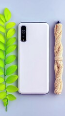 A white smartphone is placed next to a green leafy branch and a coiled rope on a light blue surface.
