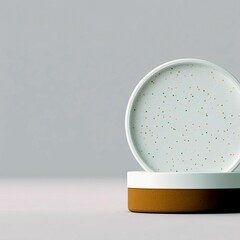A speckled plate rests on a two-toned base, creating a simple and modern display against a plain gray backdrop.